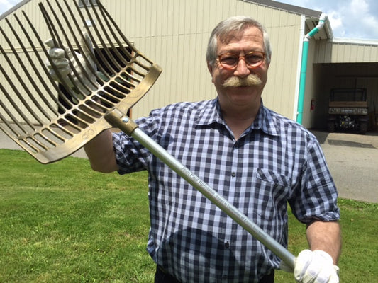A man in a plaid shirt and gloves holds up a large metal rake outdoors, perhaps searching for an Expired Forever Fork from Forever Fork, with grass and a beige building in the background.