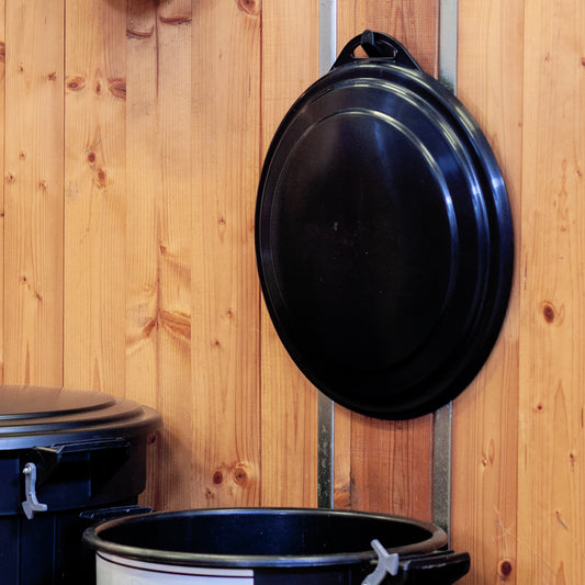 A black plastic garbage can lid hangs on a Plastic 2" Stable Hook by Forever Fork, which is attached to a wooden wall next to a black trash bin with its lid open.