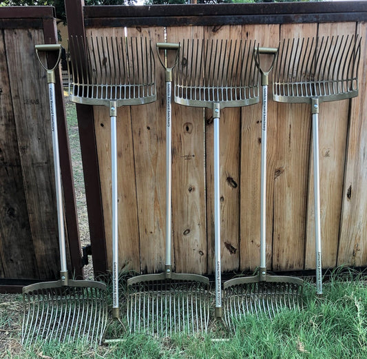 Six Forever Fork brand garden forks from the Big Barn set are lined up vertically against a wooden fence on grass.